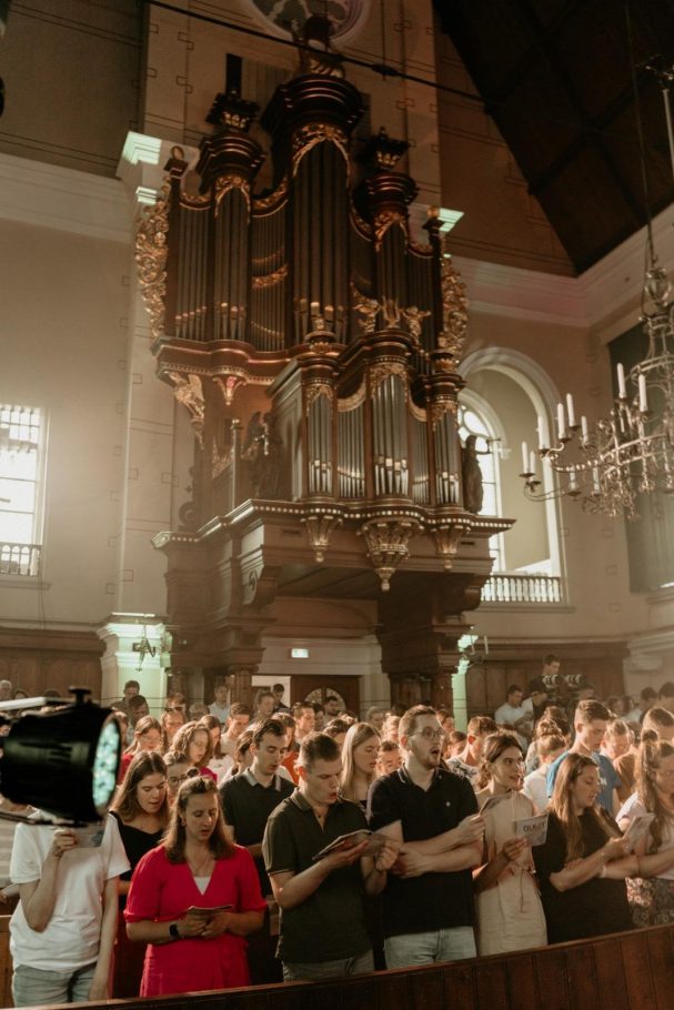 Een volle kerk met mensen die muziek zingen, achter een groot orgel.